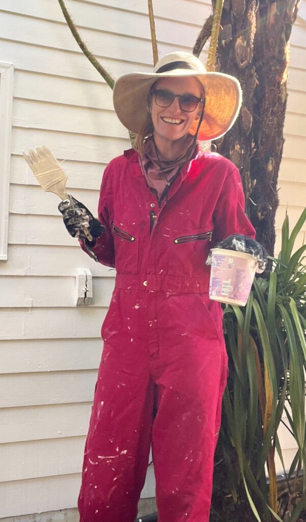 Cece in red coveralls with white paint splatters, smiling while holding a paintbrush in front of a freshly painted wall in Sonoma County.